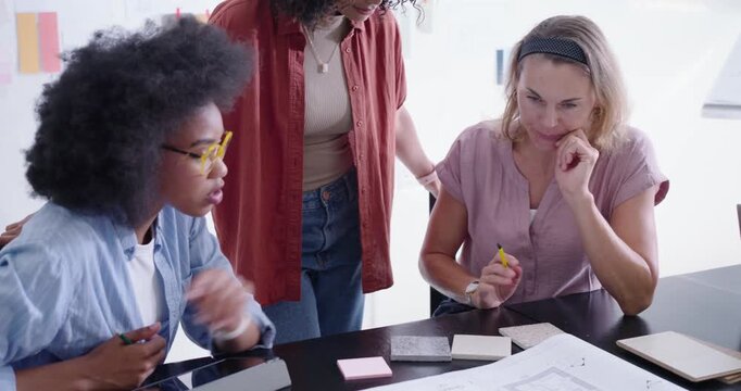 Three women starting studio review, leaning over blueprint, selecting finishes with yellow pencil