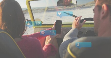 Fototapeta premium Driving couple in yellow drop-top along beach, passenger in pink sweater holding phone with badges