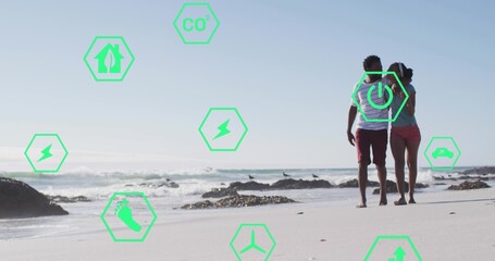 Fototapeta premium Walking couple wearing casual beachwear along wet sand, green hex overlays with icons, copy space