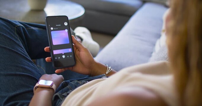 African American woman on couch holding phone studying purple card and tapping, smartwatch on lap