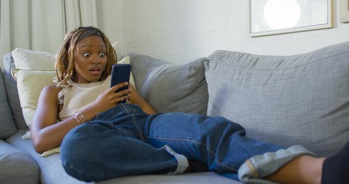 Adult African woman lying on sofa at home reading shocking smartphone message, reacting with worry