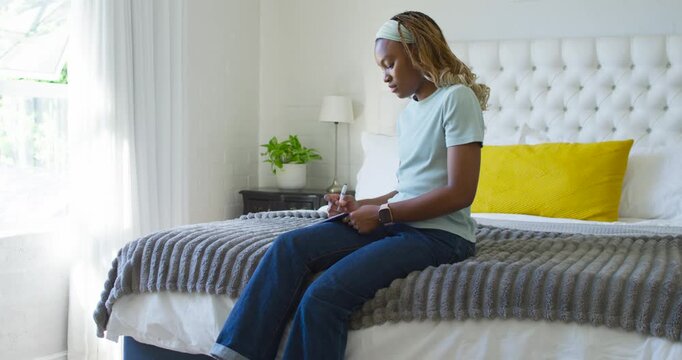 Adult African American woman writing in notebook with pen on bed, pausing, reflecting in sunlight