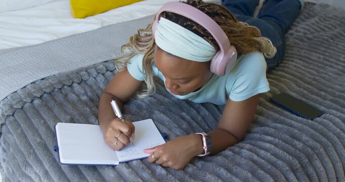 African American woman lying on bed with pink headphones using phone while taking notes in notebook