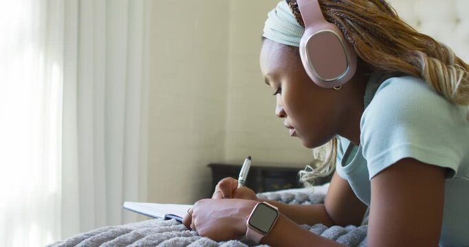 African female student lying on bed by window, listening on headphones and taking notes in notebook