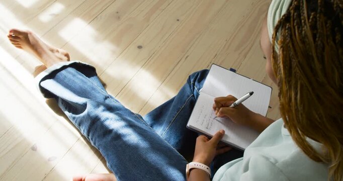 African American woman sitting on plank floor placing notebook and journaling while sunlight shifts
