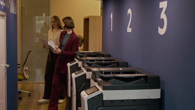 Young businesswoman talking with female colleague, then walking to multifunction printer and copying documents during office workday