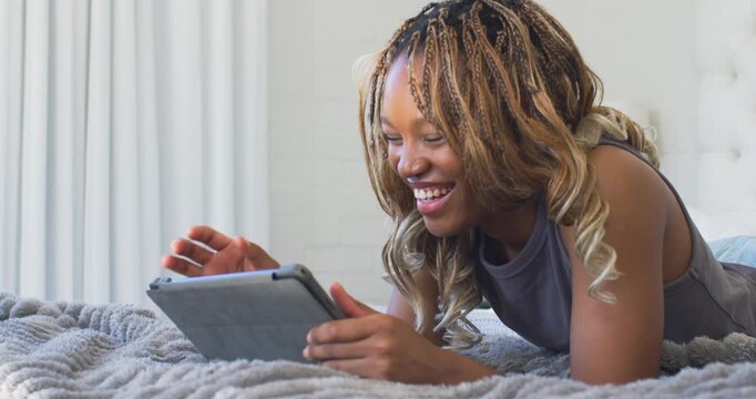 African American woman lying on bed atop plush blanket holding tablet, tapping, laughing at content
