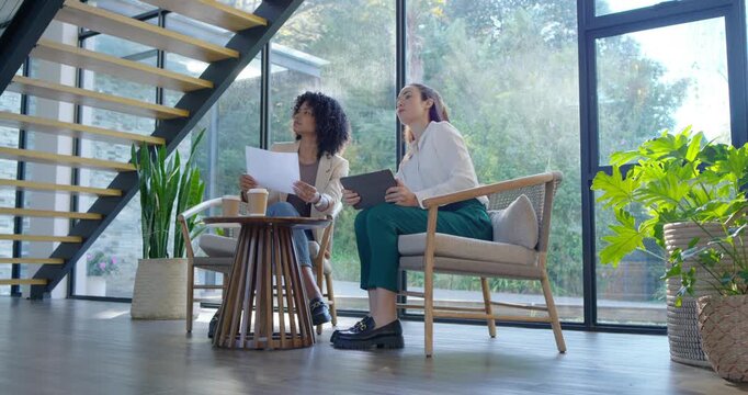 Diverse female coworkers checking tablet, comparing papers, pointing to staircase design in lounge