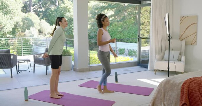 African American woman cueing from tablet, Asian woman following and both warming up on purple mats