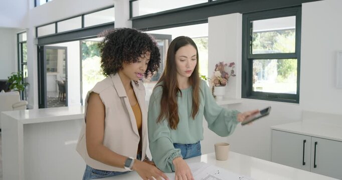 Diverse female team spreading blueprint at kitchen island pointing and using tablet finalizing plan
