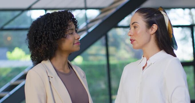 Diverse female coworkers starting work talk, right speaking while left in blazer nodding in atrium