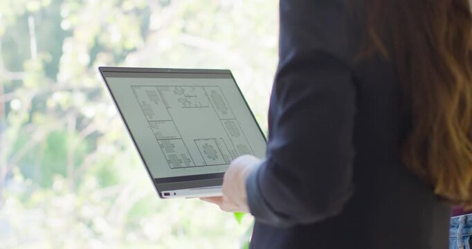 Adult female in dark blazer holding laptop by window tilting, showing layout for camera, copy space