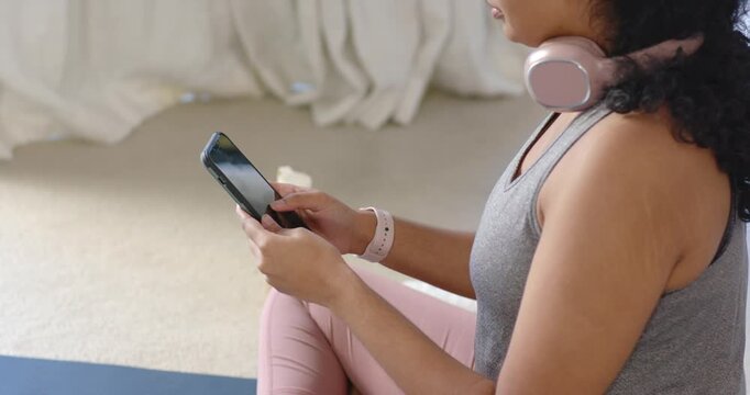 Adult woman sitting on yoga mat in bedroom tapping phone, listening via pink headphones, copy space