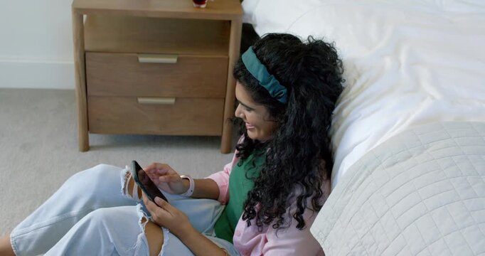 Female sitting on floor by bed, bringing smartphone up, scrolling and tapping messages, smiling