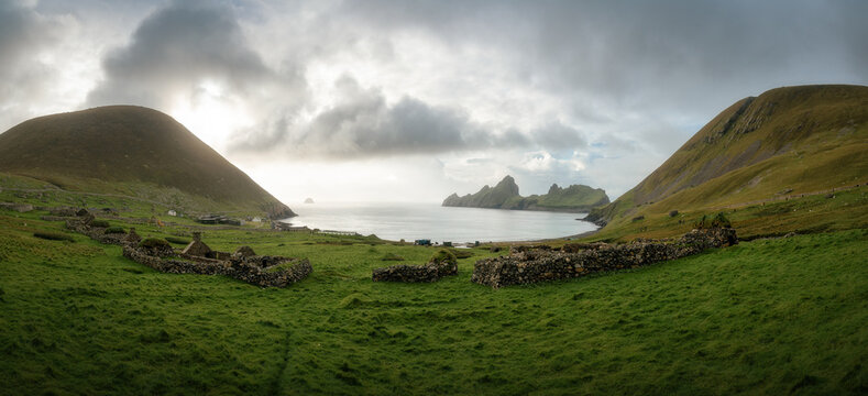 View of the historic stone ruins on Hirta island with green rolling hills and the sea bay under a cloudy sky in Scotland, United Kingdom.