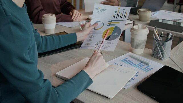 Close view of businesswoman holding printed charts, analyzing financial data and taking notes in notebook at office desk