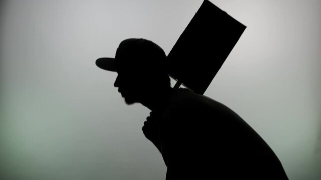 An anonymous silhouette of a man in a cap, dragging a poster behind his back against a white wall