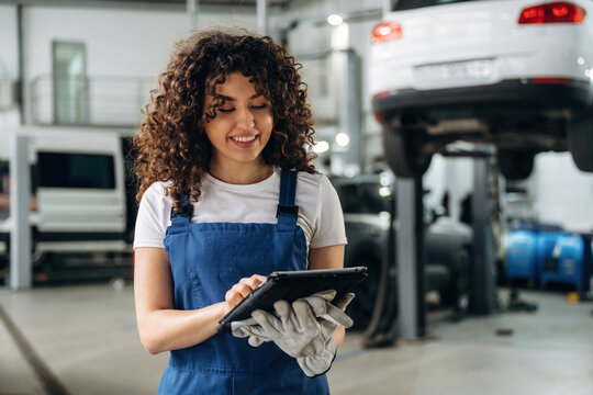 Female auto mechanic with curly hair using tablet in garage, vehicles elevated on lifts, tools and equipment visible in the background