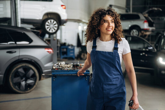 Female auto mechanic with curly hair wearing blue overalls smiles while holding a tool in a busy automotive garage with cars in the background