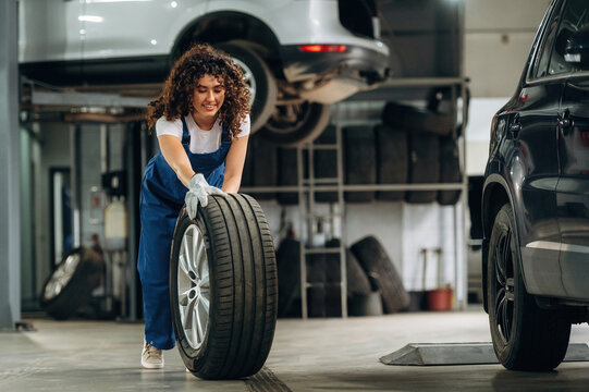 Female auto mechanic with curly hair lifts a car tire inside a garage, with vehicles elevated on lifts and stacks of tires visible in the background