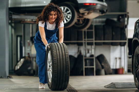 Female auto mechanic with curly hair lifts a large tire inside a garage, with vehicles elevated on lifts and tools visible in the background