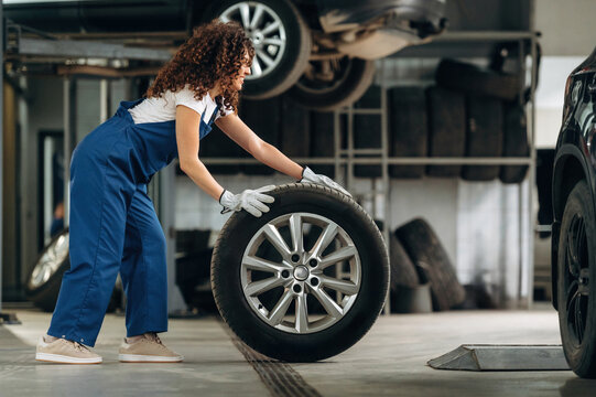 Female auto mechanic in blue overalls lifts a tire inside a garage with vehicles elevated on lifts and tools visible in the background