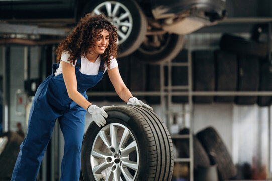 Female auto mechanic with curly hair lifts a tire in a garage filled with car wheels and tools, showcasing a busy automotive workshop environment