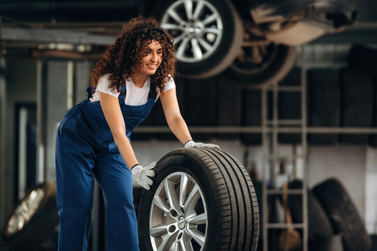 Female auto mechanic with curly hair lifts a car tire in a garage, surrounded by shelves of tires and automotive equipment in the background
