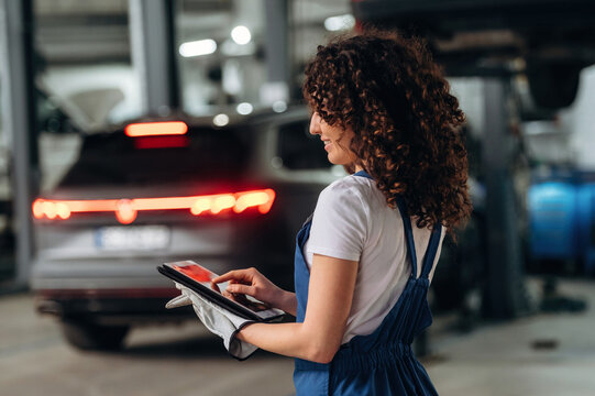 Female auto mechanic with curly hair using tablet to check vehicle diagnostics in a modern garage with cars and tools in the background