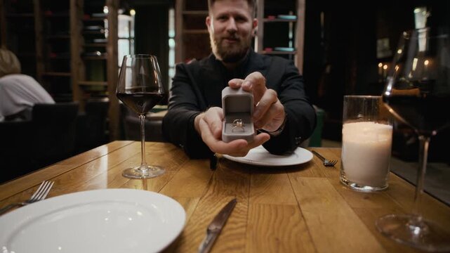 POV handheld shot of young Caucasian man in formalwear sitting at table in luxury restaurant presenting engagement ring in jewelry box, proposing marriage to unrecognizable woman saying yes