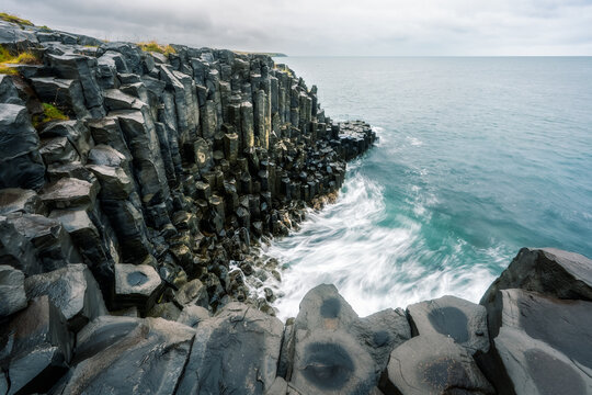 View of basalt columns along the rugged coastline with waves crashing against the dark rocks under a cloudy sky in Hofsos, Skagafjordur, Iceland.