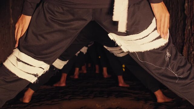 From a low-angle viewpoint, a group of contemporary dancers in dark, tribal costumes performs a synchronized routine in a dimly lit, narrow passage. A mysterious and artistic performance.