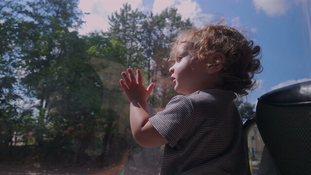 Toddler presses hand and face against bus window while observing outdoors, immersed in changing scenery with blue sky and trees reflected on glass
