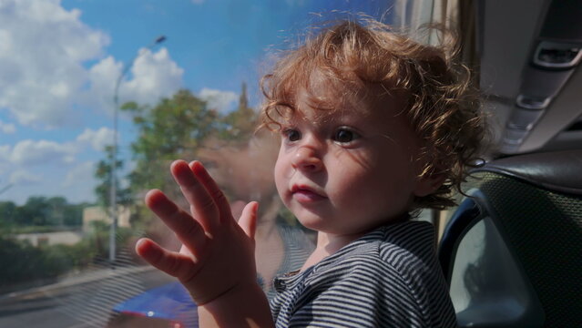 Toddler gazes through bus window with hand pressed to glass, face half-lit by sun and surrounded by blue sky and fluffy clouds reflected on surface