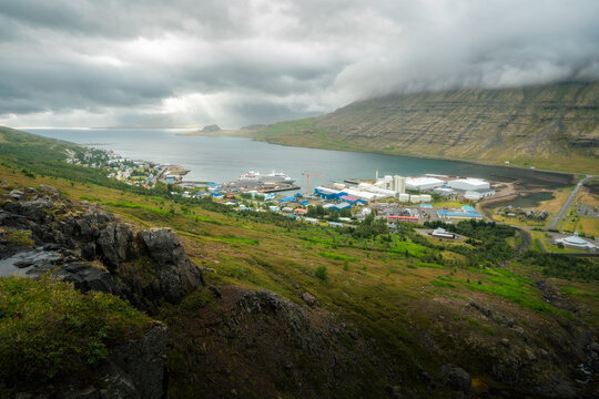 View of the coastal town of Eskifjordur in a fjord with a cruise ship docked at the harbor under a dramatic cloudy sky with sunbeams hitting the water Eskifjordur, Fjardabyggd, Iceland.