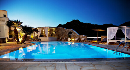 Luxury swimming pool at night with soft ambient lighting and reflections on water. Mediterranean resort setting with stone architecture, palm trees and relaxing atmosphere. © Nikos