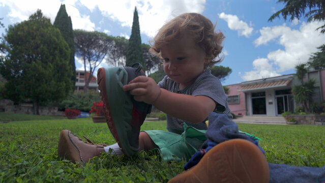 Curly haired toddler attempts to put on sock over shoe while sitting on grass in front of building, practicing dressing motions outdoors on sunny day