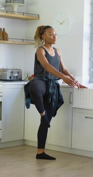 Vertical video: African American woman starting leg extensions in kitchen using counter for balance