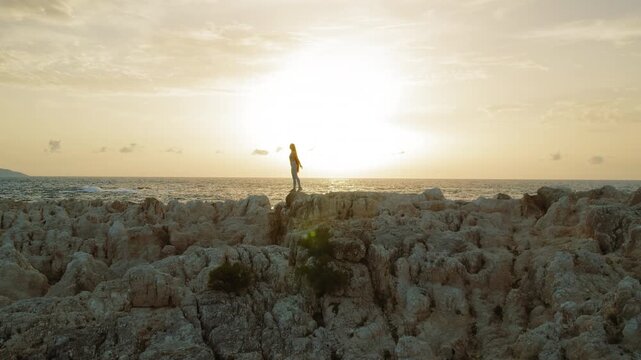 Lone woman figure stands atop coastal rocks silhouetted against a golden sunset