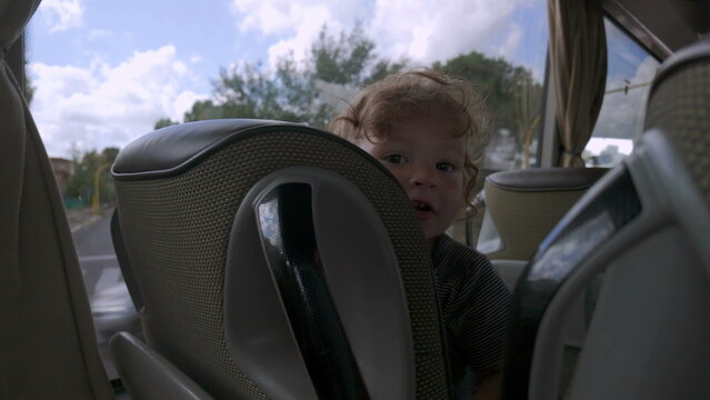 Toddler peeks between bus seats with playful expression, smiling slightly while wedged in small space during public transportation ride