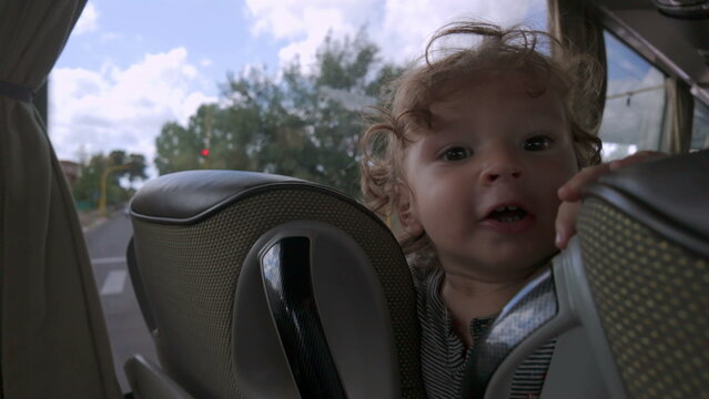 Toddler peeks between bus seats with playful expression, smiling slightly while wedged in small space during public transportation ride