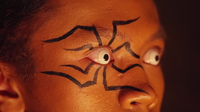 An extreme close-up on a person's wide, white-lensed eyes, surrounded by spider-leg makeup. The eyes dart around in panic and fear, creating a deeply unsettling and shocking image.