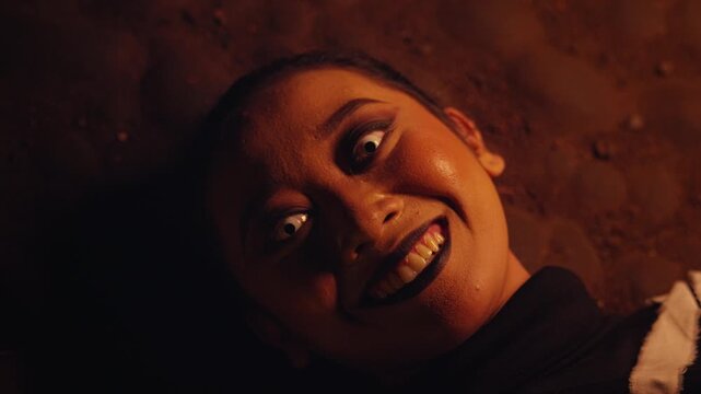 A terrifying close-up of a woman lying on the ground. She stares up with a wide, manic smile and white contact lenses, creating a deeply disturbing and insane horror portrait.