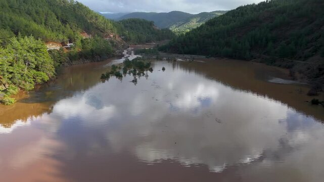 Muddy floodwaters of the Namnam River reflecting surrounding pine-forested hills in Koycegiz, Mugla, Turkey. Aerial drone view, moving forward