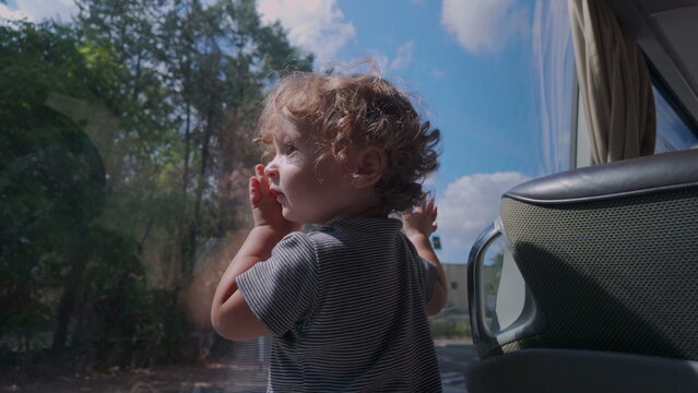 Toddler presses hand and face against bus window while observing outdoors, immersed in changing scenery with blue sky and trees reflected on glass