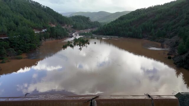 Muddy floodwaters of the Namnam River revealing surging over a concrete stepped weir surrounded by pine-forested hills in Koycegiz, Mugla, Turkey. Aerial drone view, moving backwards