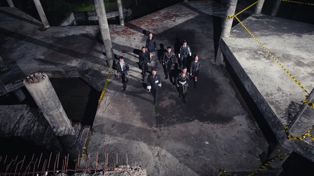 From a high angle, a group of stylishly dressed people walks through an abandoned, unfinished concrete building at night. The scene is eerie, industrial, and has a post-apocalyptic feel.