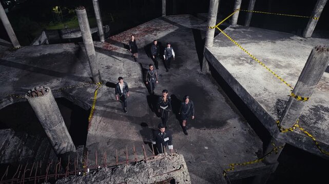 From a high-angle view, a group of people in black outfits walks through and exits a derelict, multi-level concrete structure at night. An eerie scene of departure or escape.