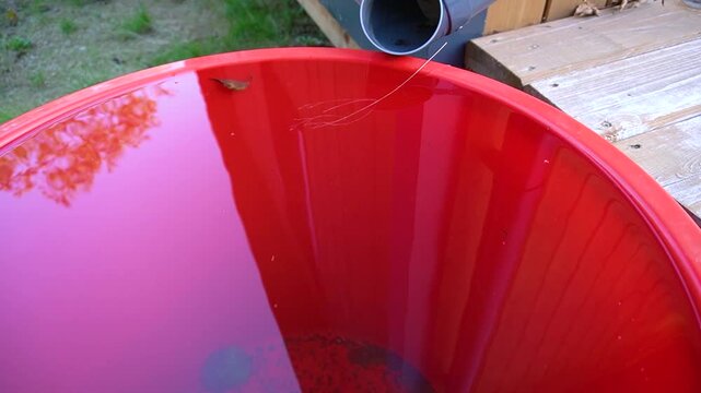 A red water barrel at the house. A plastic barrel stands under a drainpipe near a wooden private house.