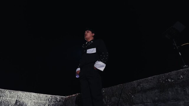 A young man in a stylish black and white outfit stands alone on a concrete ledge at night, looking around before walking down the stairs as a bright spotlight creates a dramatic flare.